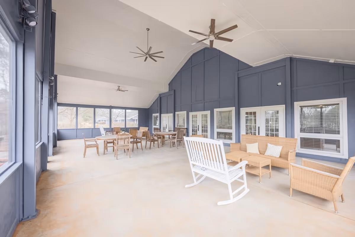 A spacious screened-in porch area with several wooden tables and chairs, a white rocking bench, and wicker seating with cushions. The walls are painted blue with multiple windows and glass doors, and the ceiling is vaulted with two large ceiling fans.
