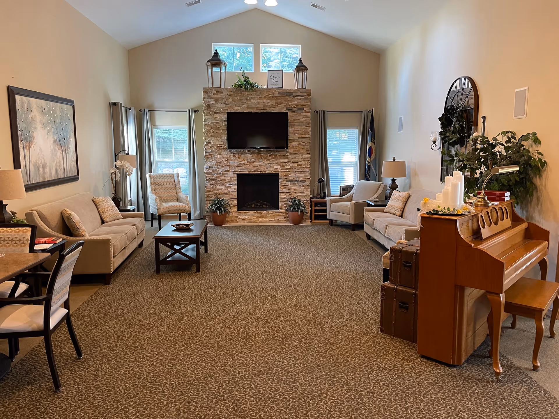 A cozy living room with beige walls and carpeted floor featuring two sofas, two armchairs, a wooden coffee table, and a stone fireplace with a mounted TV above it. There are two windows with curtains on either side of the fireplace. A wooden piano with candles and decorative items is on the right side of the room, along with some plants and a large wall clock.