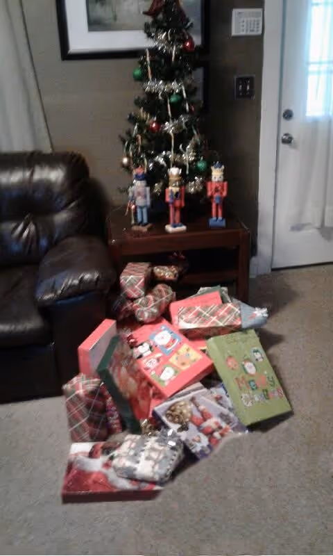 A small decorated Christmas tree on a wooden side table with three nutcracker figurines in front of it. Below the table and on the carpeted floor are numerous wrapped Christmas presents. To the left of the table is a dark brown leather armchair, and to the right is a white door with a window and curtain.