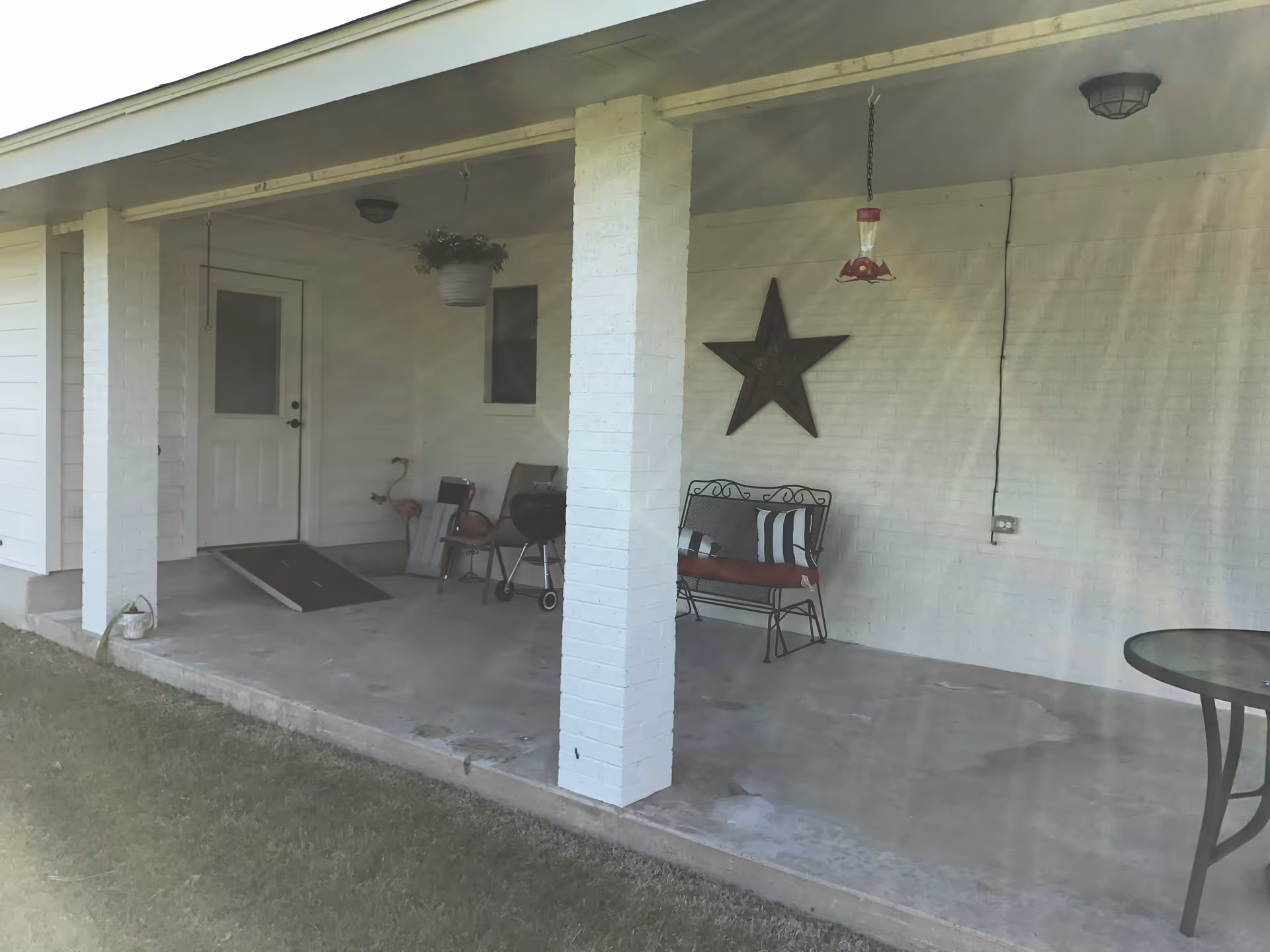 Covered outdoor patio area with white brick pillars and walls, a white door with a ramp, a hanging plant, a metal bench with cushions, a star decoration on the wall, a hanging bird feeder, a small grill, and a round glass-top table on the right side.