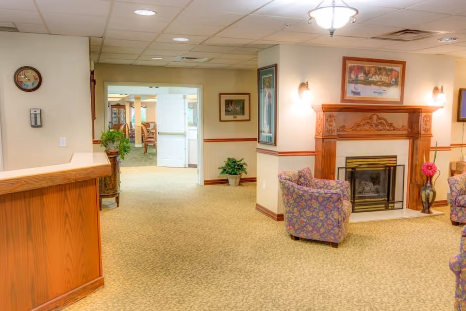 Interior view of a senior living facility common area with a wooden reception desk on the left, a floral-patterned armchair near a decorative wooden fireplace with a framed painting above it, and an open doorway leading to a dining area with tables and chairs. The walls are light-colored with wooden trim, and there are plants and framed artwork decorating the space.