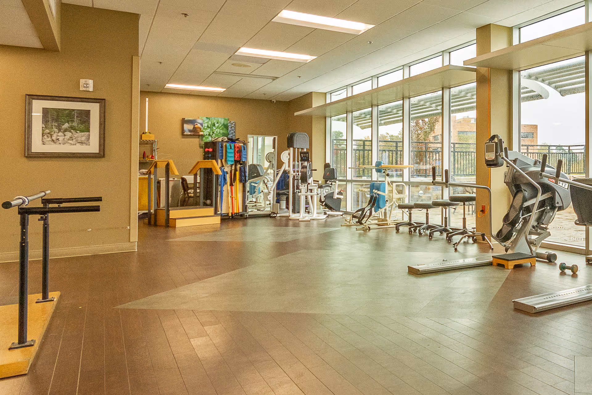 Spacious rehabilitation gym with exercise equipment, parallel bars, and floor-to-ceiling windows.
