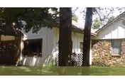 Front exterior of a single-story building with white siding, stone wall, and trees.