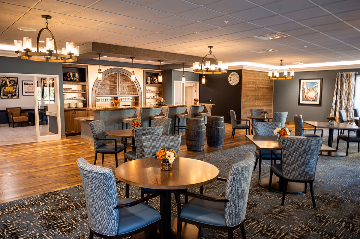 Spacious dining and common area with round tables, upholstered chairs, a bar counter and warm overhead lighting.