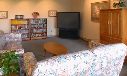 A cozy living room with floral patterned sofas arranged around a wooden oval coffee table. There is a large flat-screen TV on a stand against the wall, a wooden bookshelf filled with books, and a wooden cabinet with a framed picture and flowers on top. The room has soft lighting and a carpeted floor.