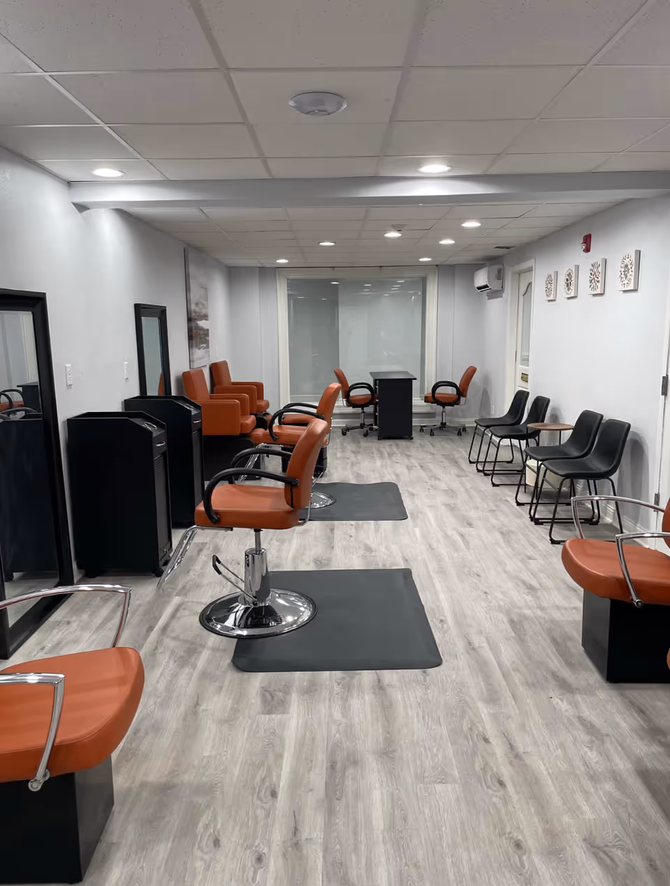 Interior view of a salon area with multiple orange salon chairs and mirrors on the left side, black chairs lined up against the right wall, light wood flooring, and a small desk with two chairs at the far end of the room.