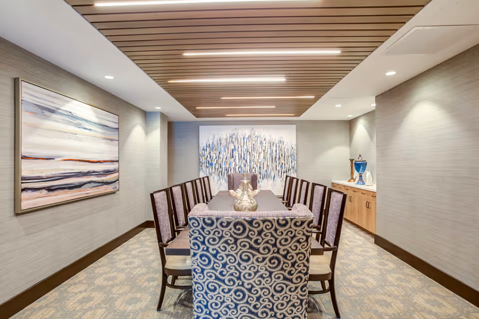 A modern dining room with a long rectangular table surrounded by ten chairs, including two patterned armchairs at the ends. The room features a wooden slatted ceiling with recessed lighting, neutral-colored walls with two large abstract paintings, and a sideboard with decorative items on the right side.