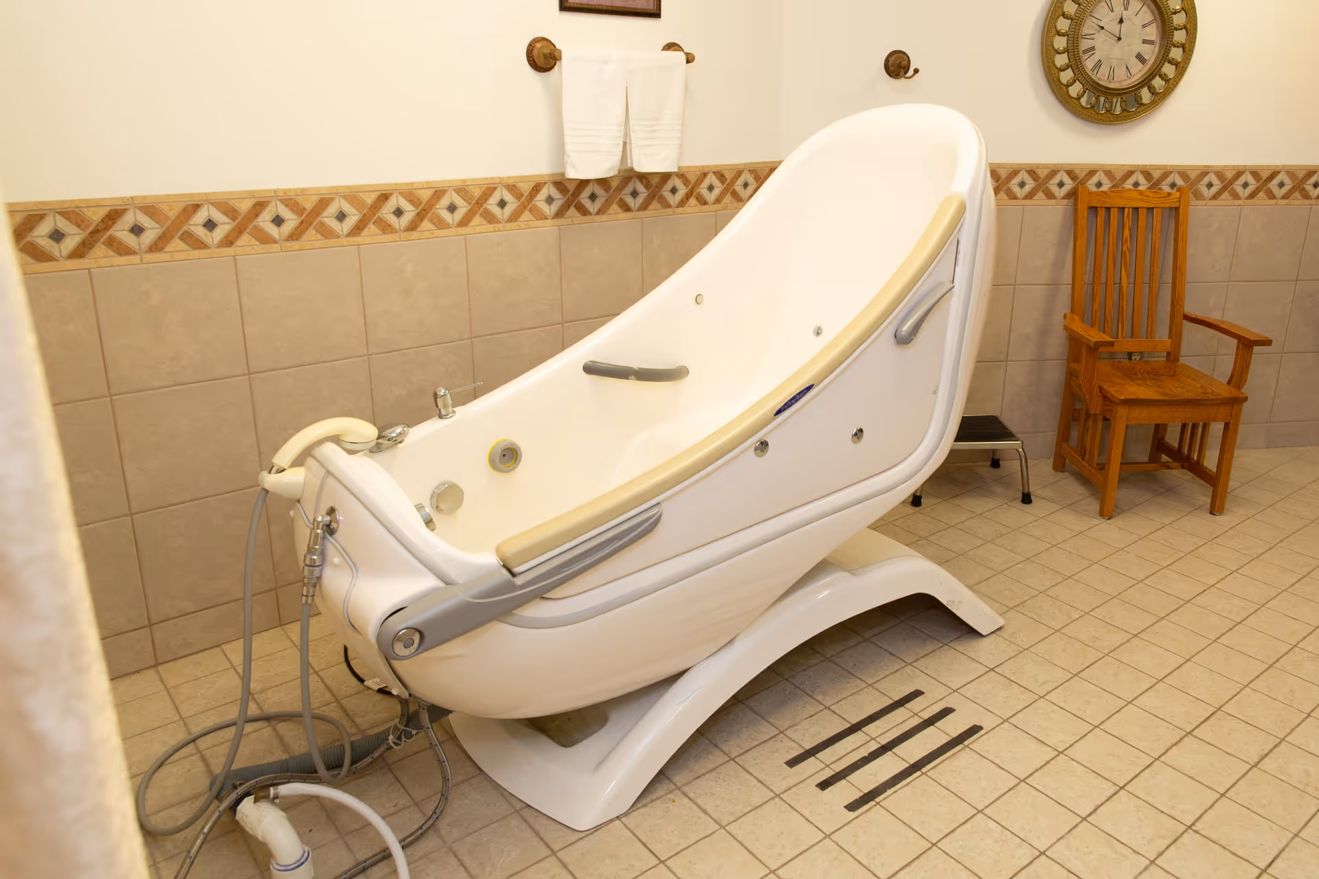 An assisted bathing tub in a tiled bathroom with a wooden chair and wall clock on the wall.