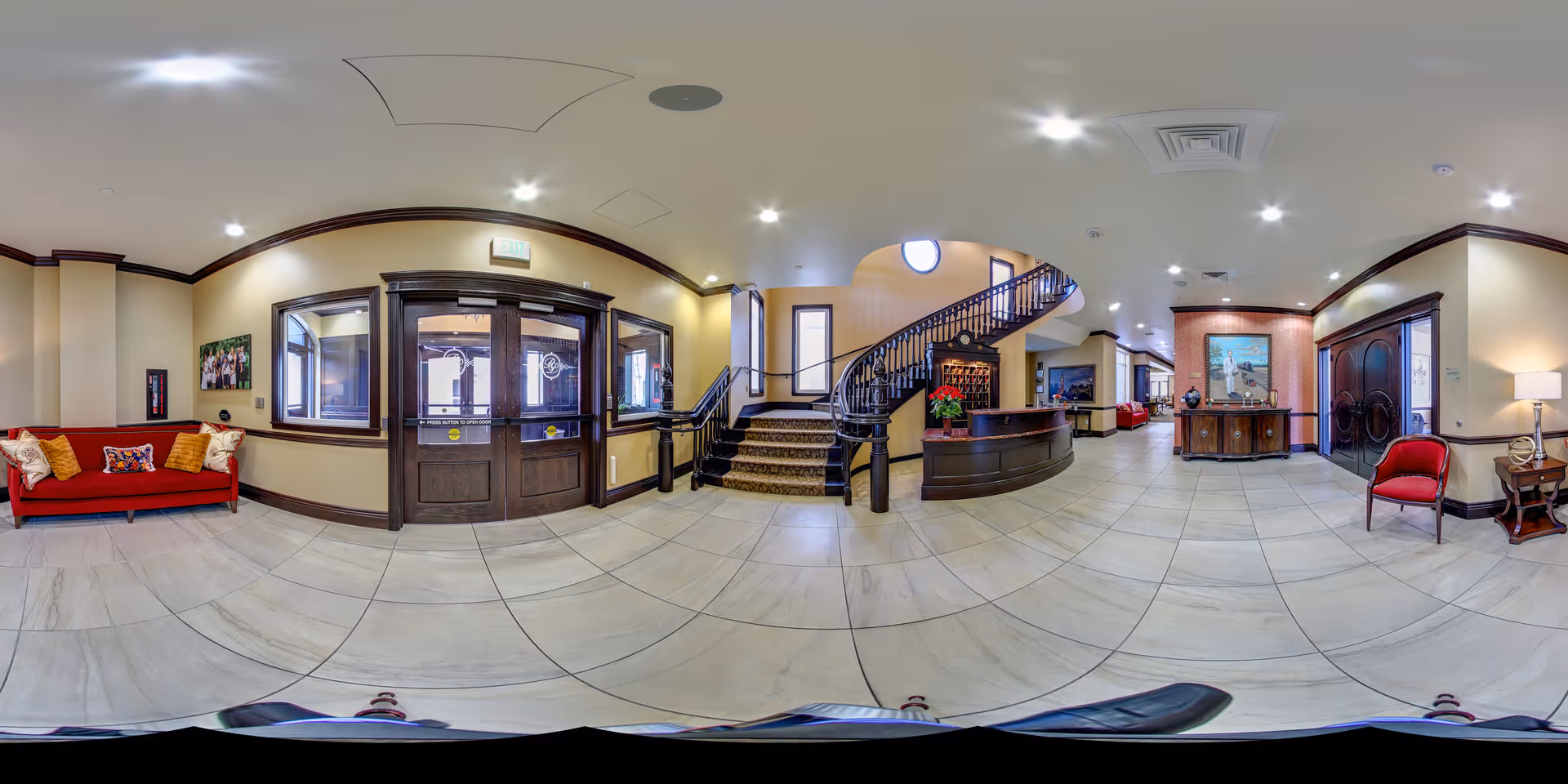 A spacious and well-lit senior living facility lobby with beige tiled floors and cream-colored walls. The area features a red sofa with decorative pillows on the left, a dark wooden reception desk in the center near a curved staircase with a carpet runner, and a red armchair with a side table and lamp on the right. There are large wooden double doors, windows, and framed artwork on the walls, creating a welcoming atmosphere.