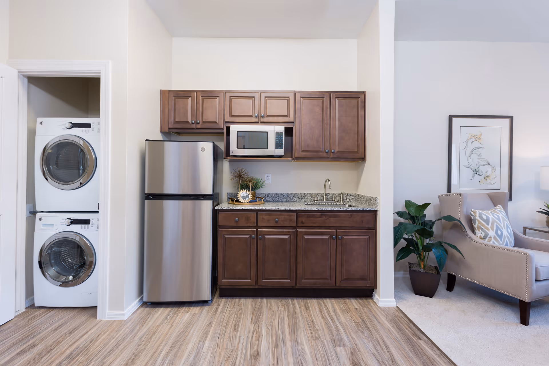 A compact kitchen area with dark wooden cabinets, a stainless steel refrigerator, a microwave, and a granite countertop with a sink. To the left, there is a stacked washer and dryer unit inside a small closet. To the right, part of a living room is visible with a beige armchair, a potted plant, and framed artwork on the wall.