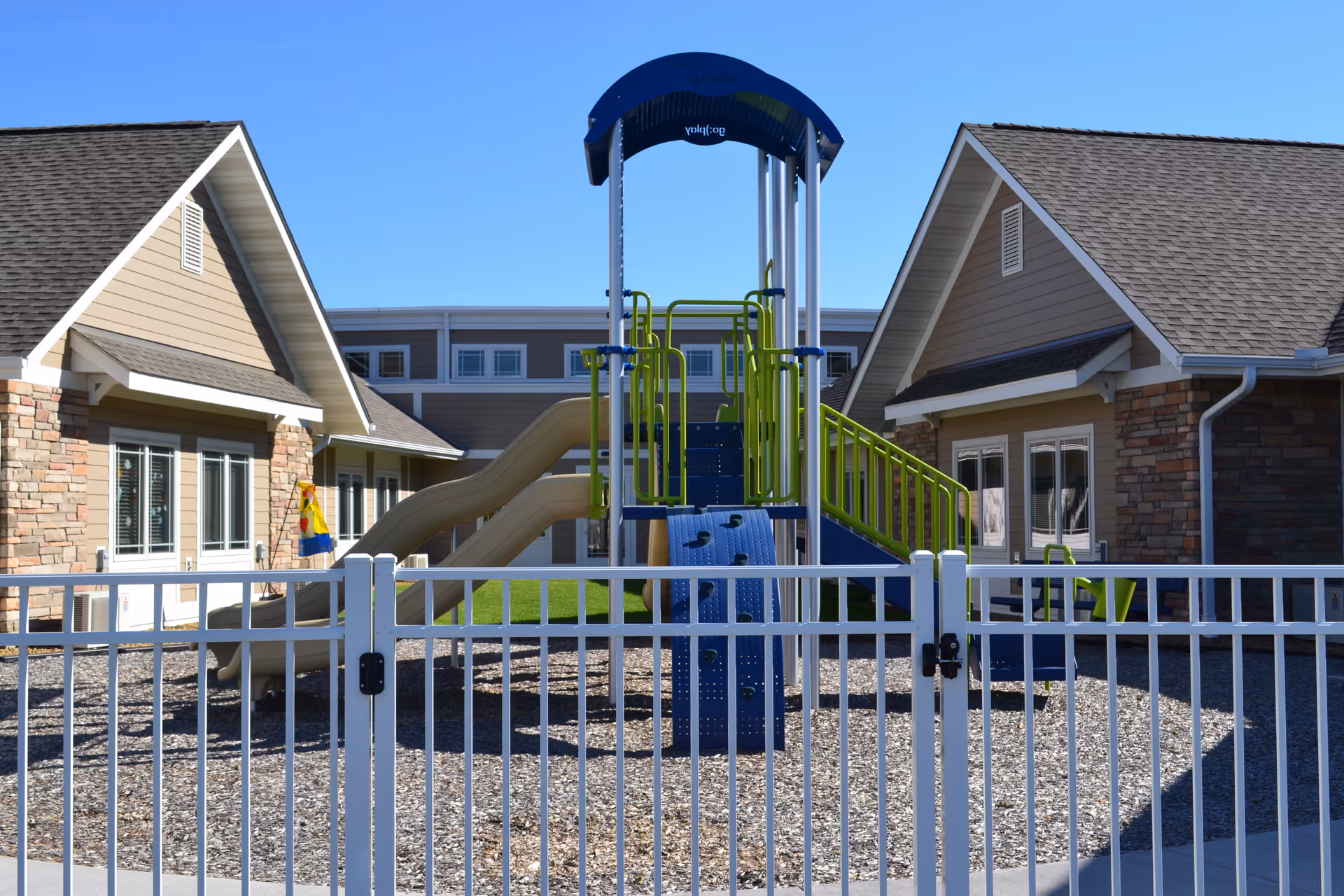 Fenced outdoor playground with slides and a climbing structure situated between two single-story facility buildings under a clear blue sky.