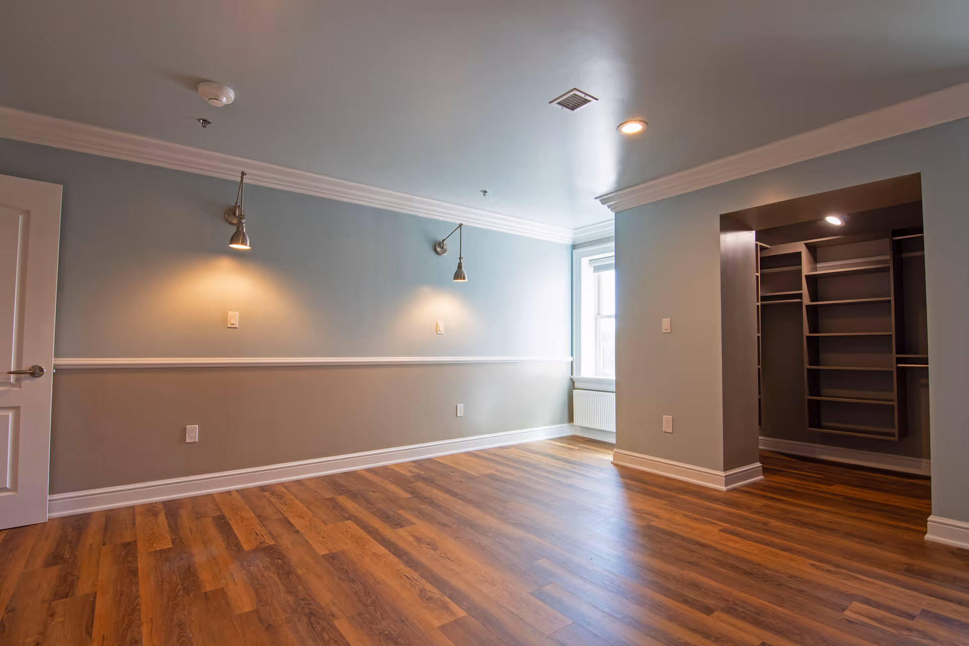 Empty room with wooden flooring, light blue and beige walls, two wall-mounted reading lights, a window letting in natural light, and an open walk-in closet with built-in shelves.