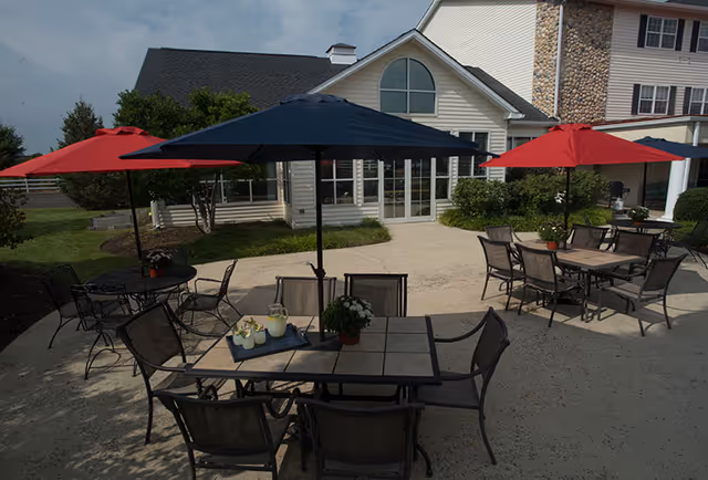 Outdoor patio area with several tables and chairs under large umbrellas in red and blue. The patio is adjacent to a building with large windows and a door, surrounded by greenery and bushes.