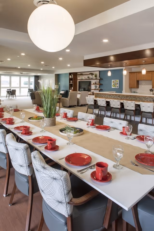 A spacious dining area with a long white table set for a meal, featuring red plates, cups, and clear glasses. The table has a beige runner and a centerpiece with tall green plants. In the background, there is a kitchen counter with bar stools, pendant lights, and a living area with chairs and a TV mounted on a blue accent wall.