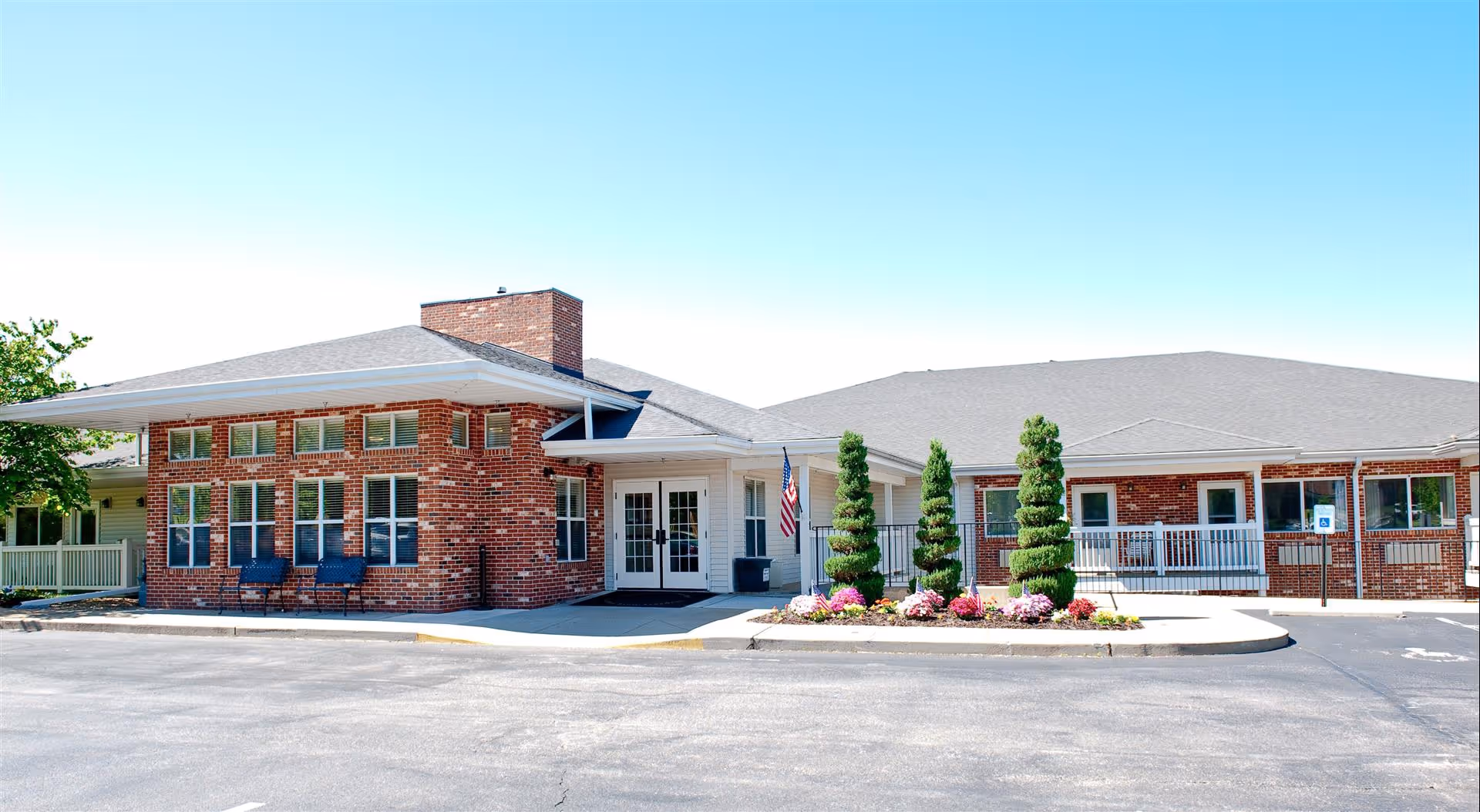 Brick single-story senior living facility front with a covered entrance, landscaped circular planter, and an American flag under a clear blue sky.