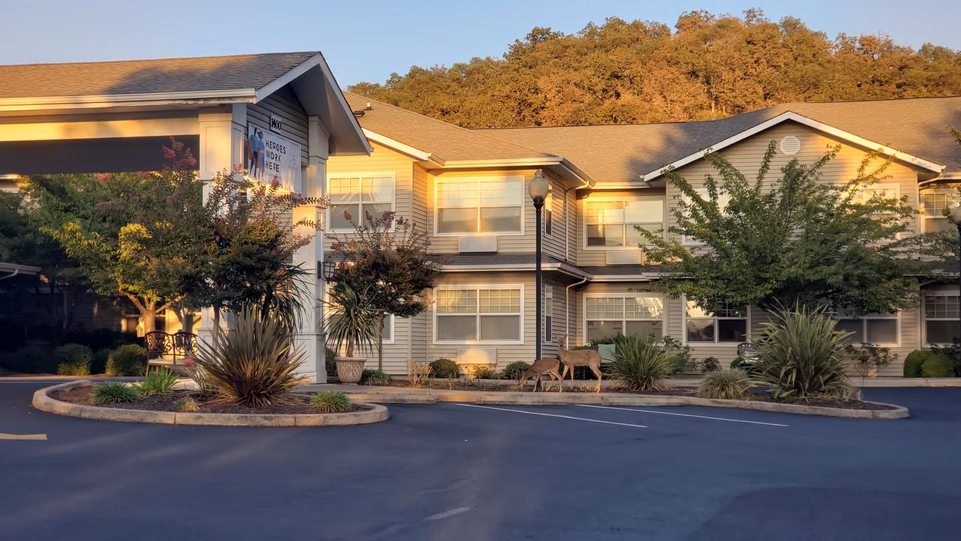Exterior view of a two-story senior living facility building with beige siding and multiple windows, surrounded by landscaped greenery and trees. There is a covered entrance with a sign that reads 'Heroes Work Here' and a paved parking area in front. The scene is lit by warm sunlight, and there are two deer near the landscaped area.