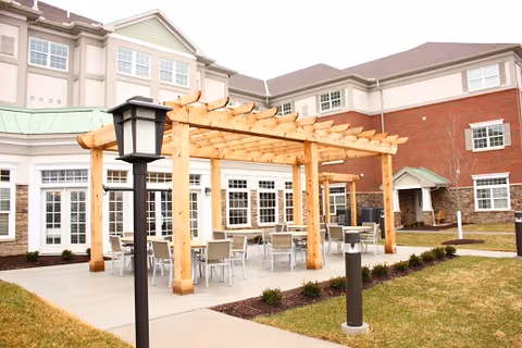Outdoor patio area at a senior living facility with a wooden pergola covering several tables and chairs. The building in the background has multiple windows and a combination of brick and light-colored siding. There are small shrubs and grass surrounding the concrete patio.