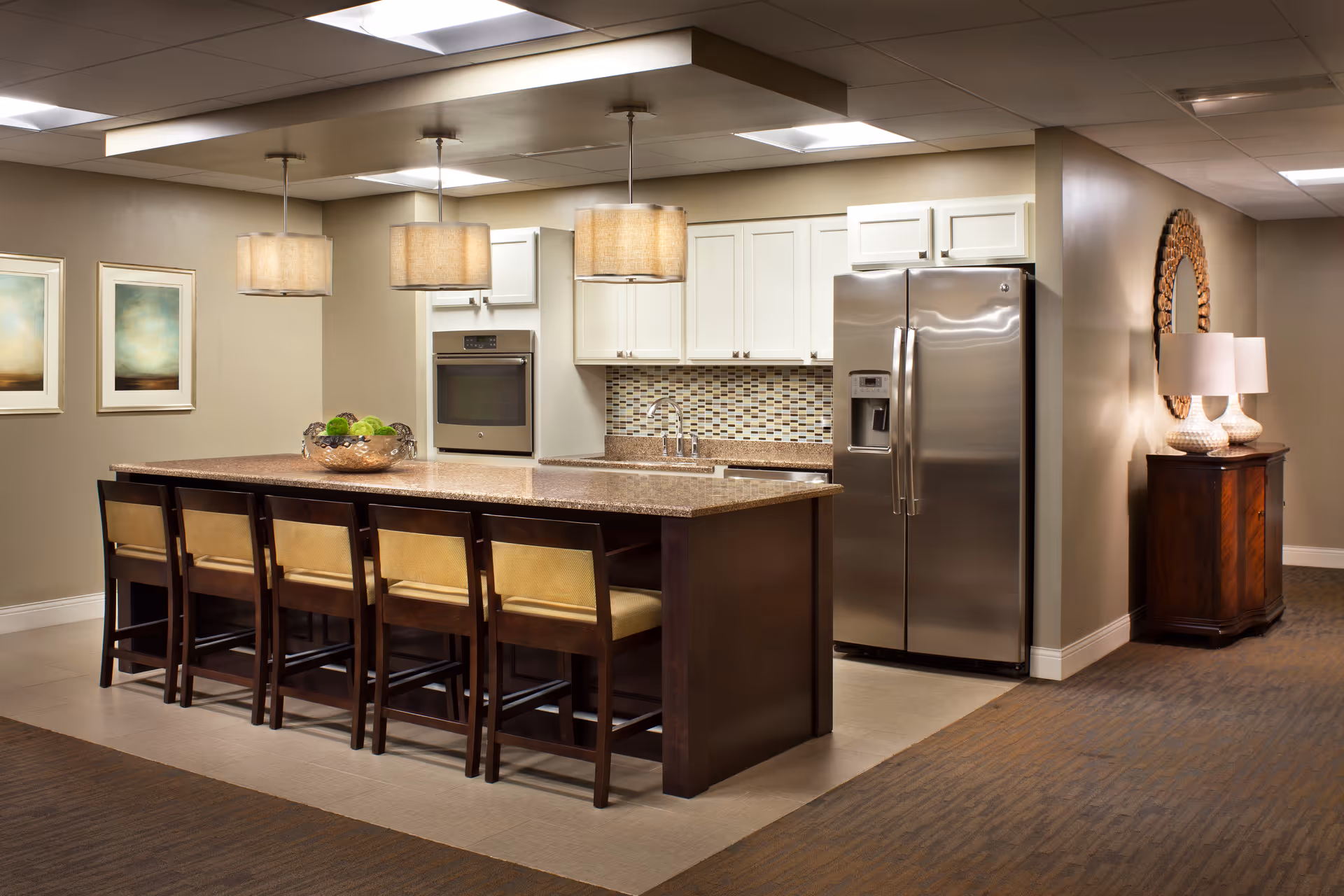 Modern communal kitchen with a long island and six bar stools, pendant lights, stainless steel refrigerator and built-in oven.