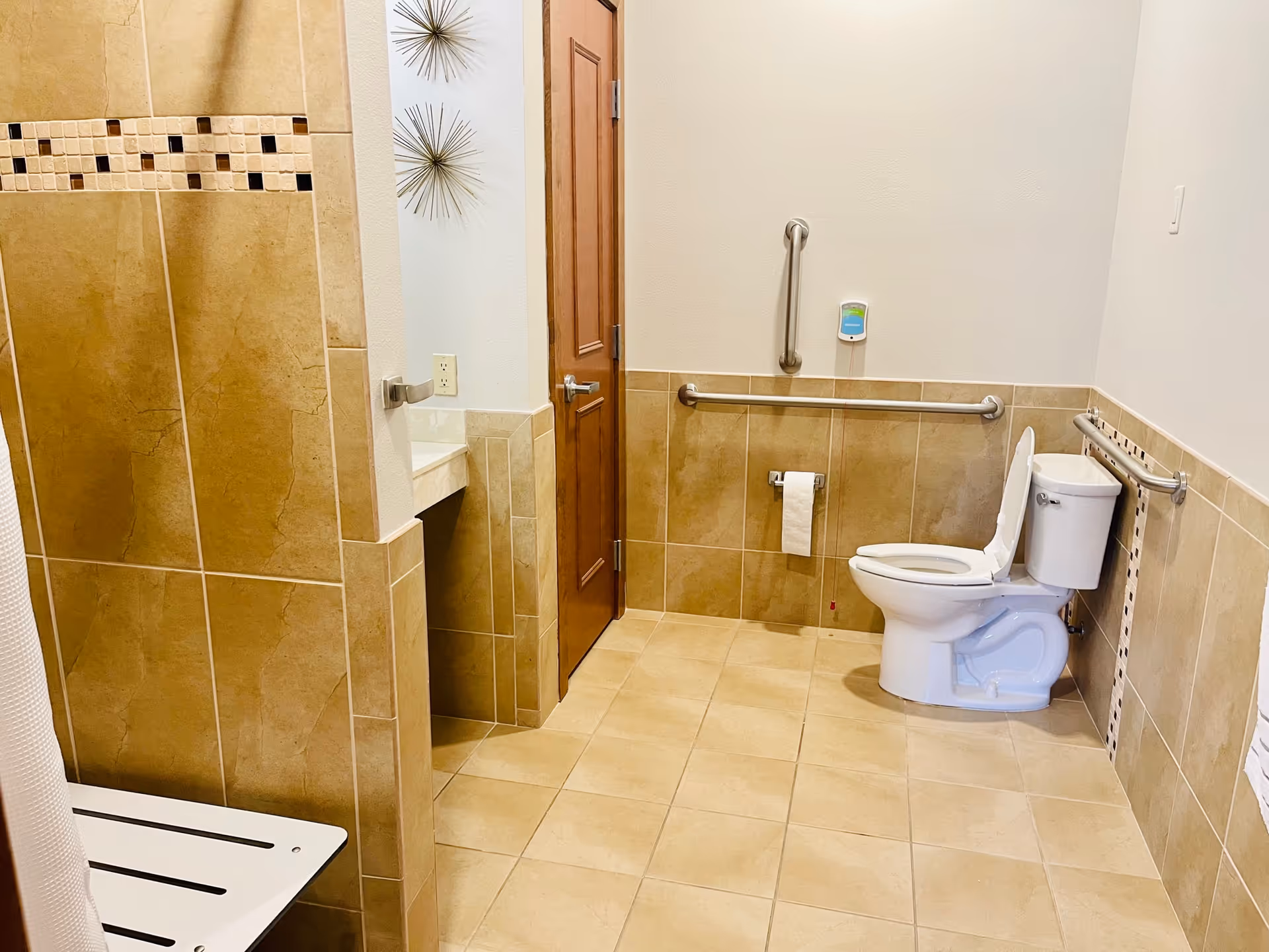Accessible tiled bathroom with a toilet, grab bars, a shower bench, and a sink area.
