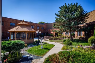 Landscaped courtyard with a wooden gazebo, paved walkways, shrubs and a surrounding brick building under a clear blue sky.