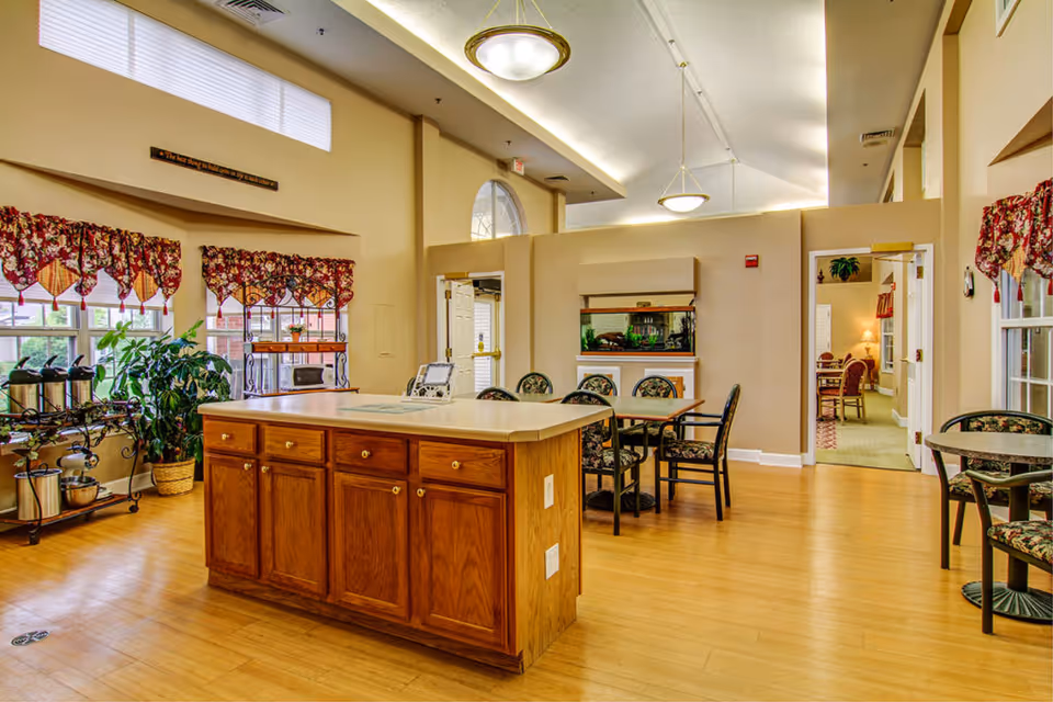 A bright and spacious common area in a senior living facility with wooden flooring, a central island with cabinets, several tables and chairs with floral upholstery, large windows with red and gold patterned valances, a plant, and an aquarium mounted on the wall.