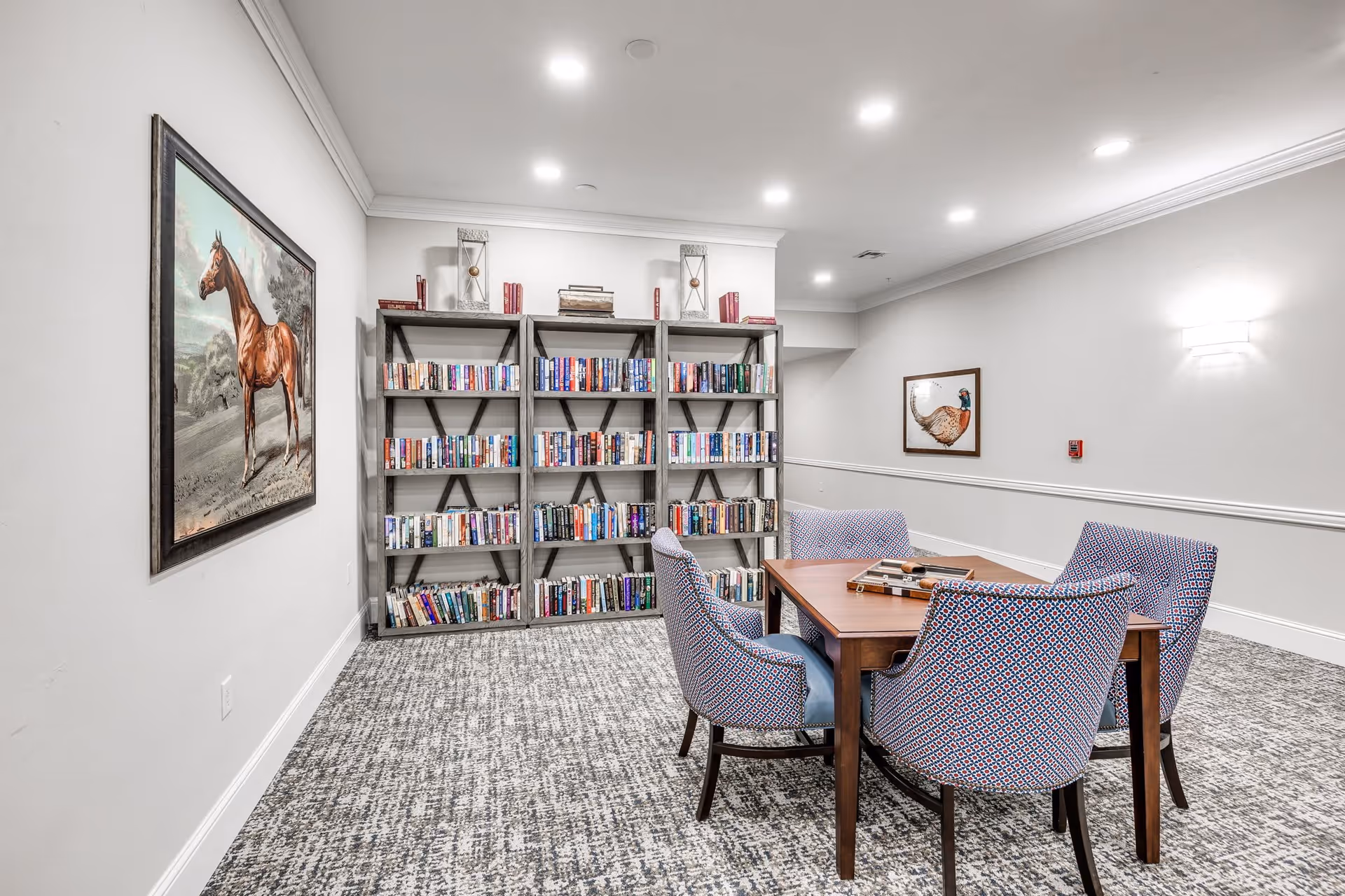 A cozy reading room in a senior living facility featuring a wooden table surrounded by four patterned upholstered chairs. Behind the table, there is a large bookshelf filled with books. The room has light-colored walls adorned with framed paintings of a horse and a bird, recessed ceiling lights, and a patterned carpeted floor.