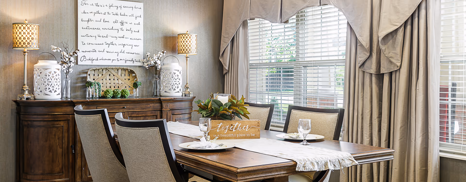 A well-lit dining room with a wooden dining table set for four, featuring beige upholstered chairs. The table has a white runner and a centerpiece with green foliage in a wooden box that reads 'together is a beautiful place to be.' Behind the table is a wooden sideboard with decorative items including two lamps with patterned shades, white lanterns, glass vases with cotton stems, and a woven basket. A large window with beige curtains and blinds lets in natural light.