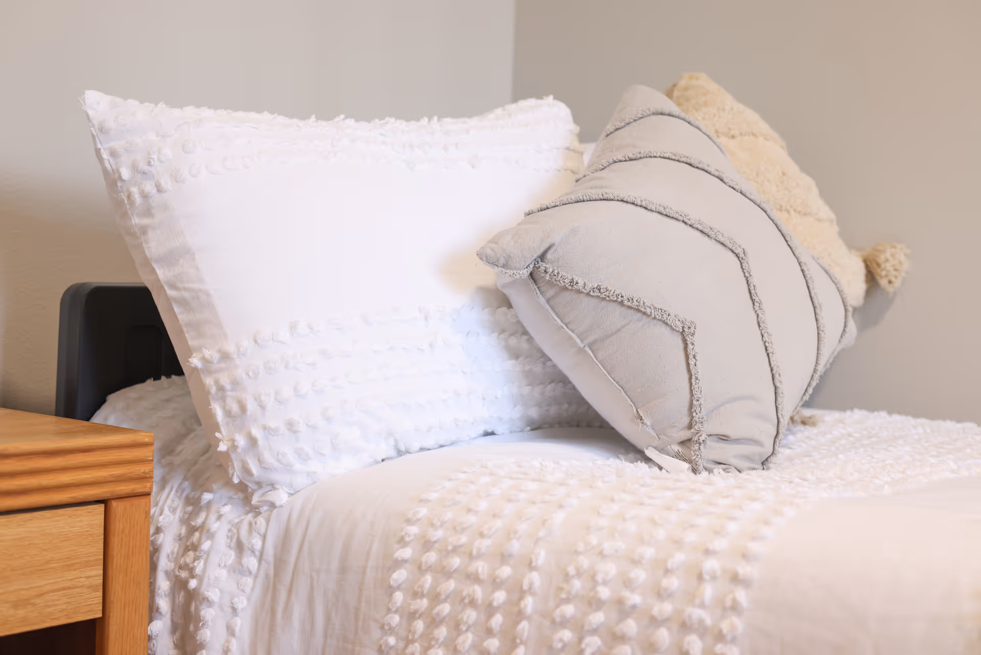 Close-up of a neatly made bed with white textured bedding, decorative pillows, and a wooden nightstand.