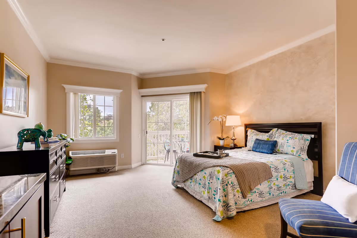 A bright and cozy bedroom featuring a queen-sized bed with floral bedding and a gray throw blanket. There is a bedside table with a lamp and a potted orchid, a blue striped chair with a white pillow, a dresser with decorative items including a green elephant figurine, and a window and sliding glass door leading to a balcony with outdoor chairs. The walls are painted beige and the floor is carpeted.