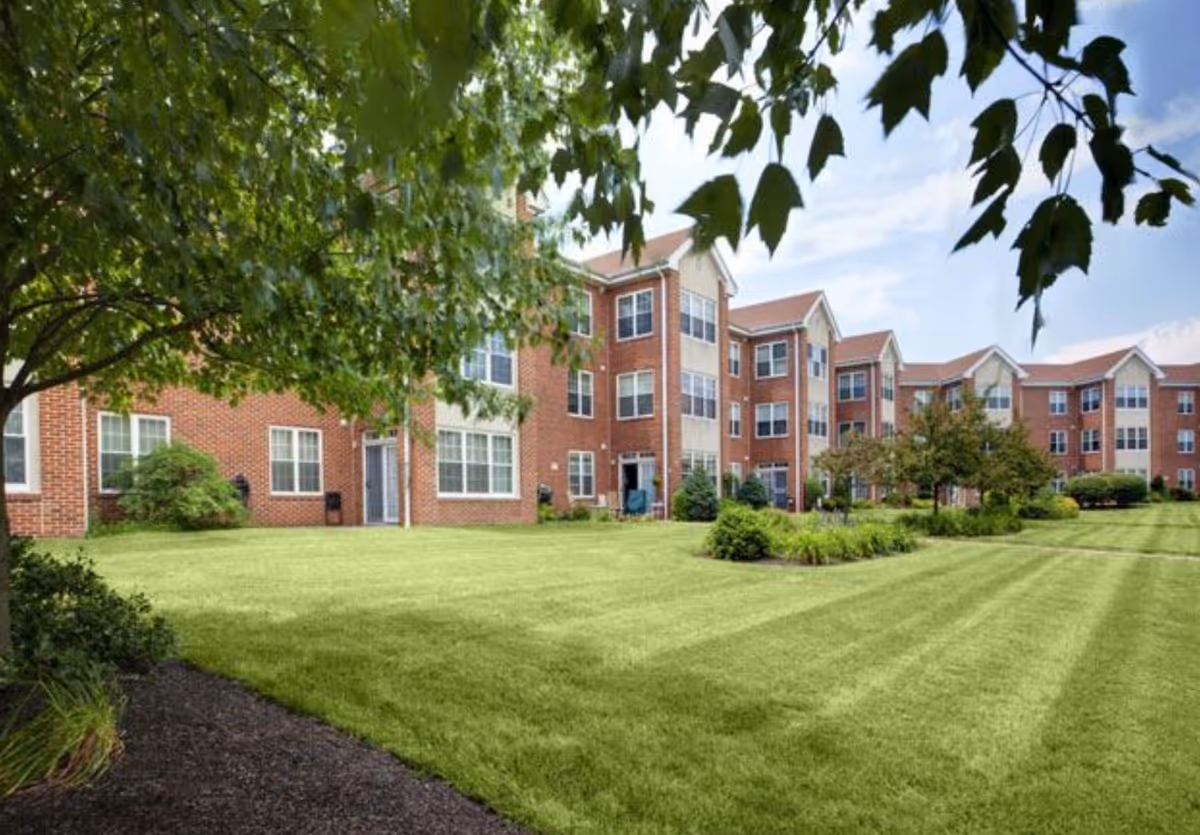 View of a large, well-maintained grassy lawn with shrubs and trees in front of a multi-story brick residential building under a partly cloudy sky.