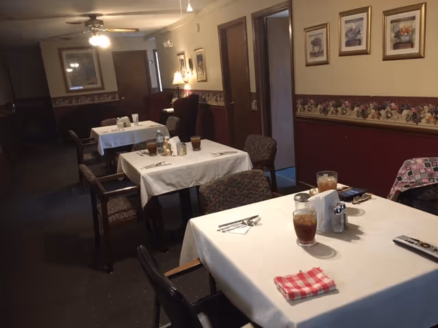 Interior view of a dining area with several tables covered in white tablecloths. Each table has chairs around it, with some tables set with glasses of iced tea, napkins, and silverware. The room has framed pictures on the walls, a ceiling fan with lights, and a lamp in the background providing warm lighting.