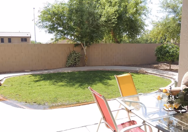 A sunny outdoor patio area with a circular patch of green grass surrounded by a concrete walkway. There are two colorful chairs, one red and one yellow, positioned around a glass table with a flower arrangement on it. Trees and bushes are visible along a beige brick wall fence in the background.