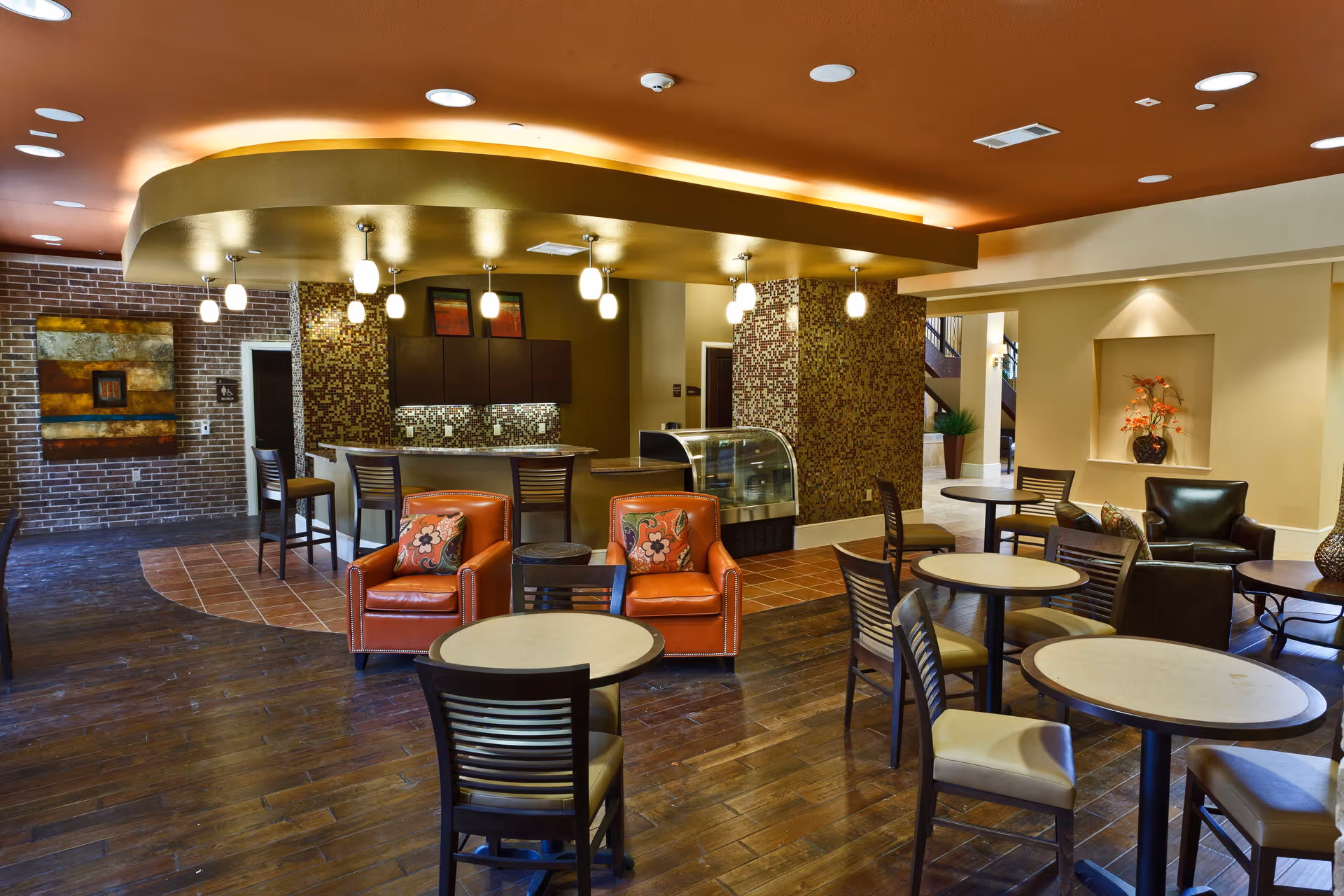 A cozy common area in Adante Independent Living featuring several round tables with chairs, two orange armchairs with floral cushions, a small bar area with high stools, pendant lighting, and decorative wall art. The space has warm lighting, wooden flooring, and a mix of brick and mosaic tile walls.