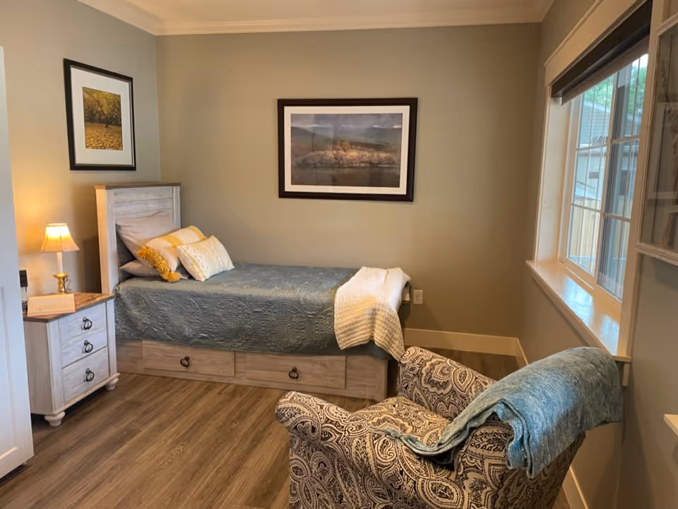 A cozy bedroom with a single bed covered in a blue quilt and decorative pillows, a wooden nightstand with a lamp, a patterned armchair with a blue throw blanket, two framed pictures on the walls, and a window letting in natural light.