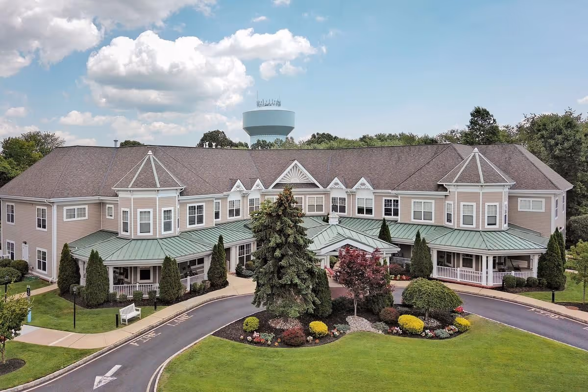 Exterior view of a large two-story senior living facility building with beige siding and a green metal roof over the entrance. The building is surrounded by well-maintained landscaping including trees, bushes, and colorful flowers. A curved driveway leads to the entrance under a partly cloudy sky.