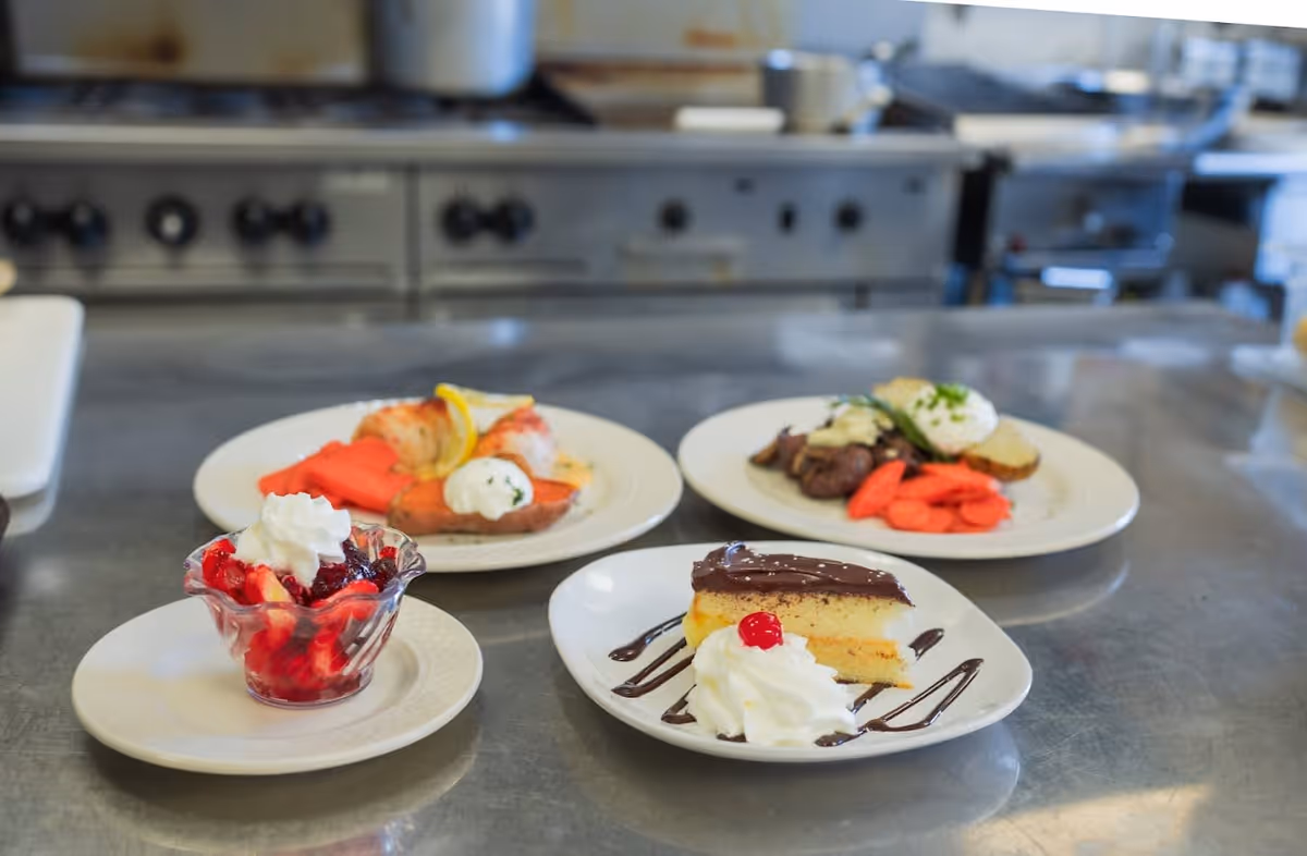 Four plates of food on a metal kitchen counter. The plates include a dessert with whipped cream and a cherry, a fruit cup with whipped cream, and two main dishes with vegetables and garnishes. The background shows a commercial kitchen stove and other kitchen equipment.
