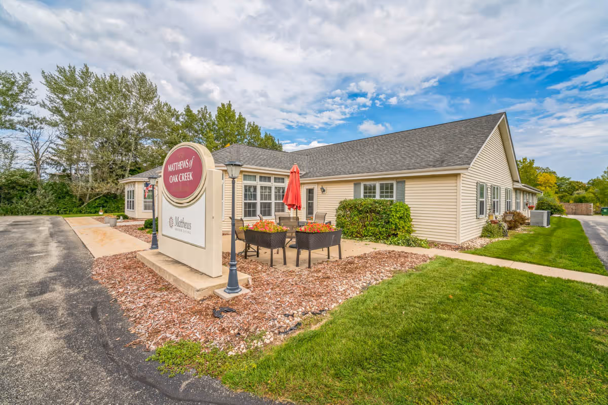 Single-story beige senior living building with a front sign, patio seating with a red umbrella, and landscaped grounds under a partly cloudy sky.