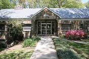 Front exterior view of a single-story nursing home building with a stone facade, a central entrance with glass doors, surrounded by greenery including bushes and trees, and a concrete walkway leading to the entrance.