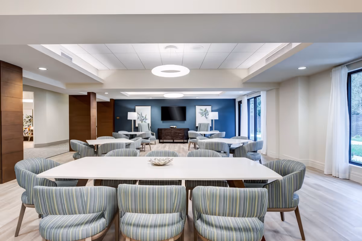Bright communal dining and lounge area with white tables, striped upholstered chairs, seating groups, and a wall-mounted TV.