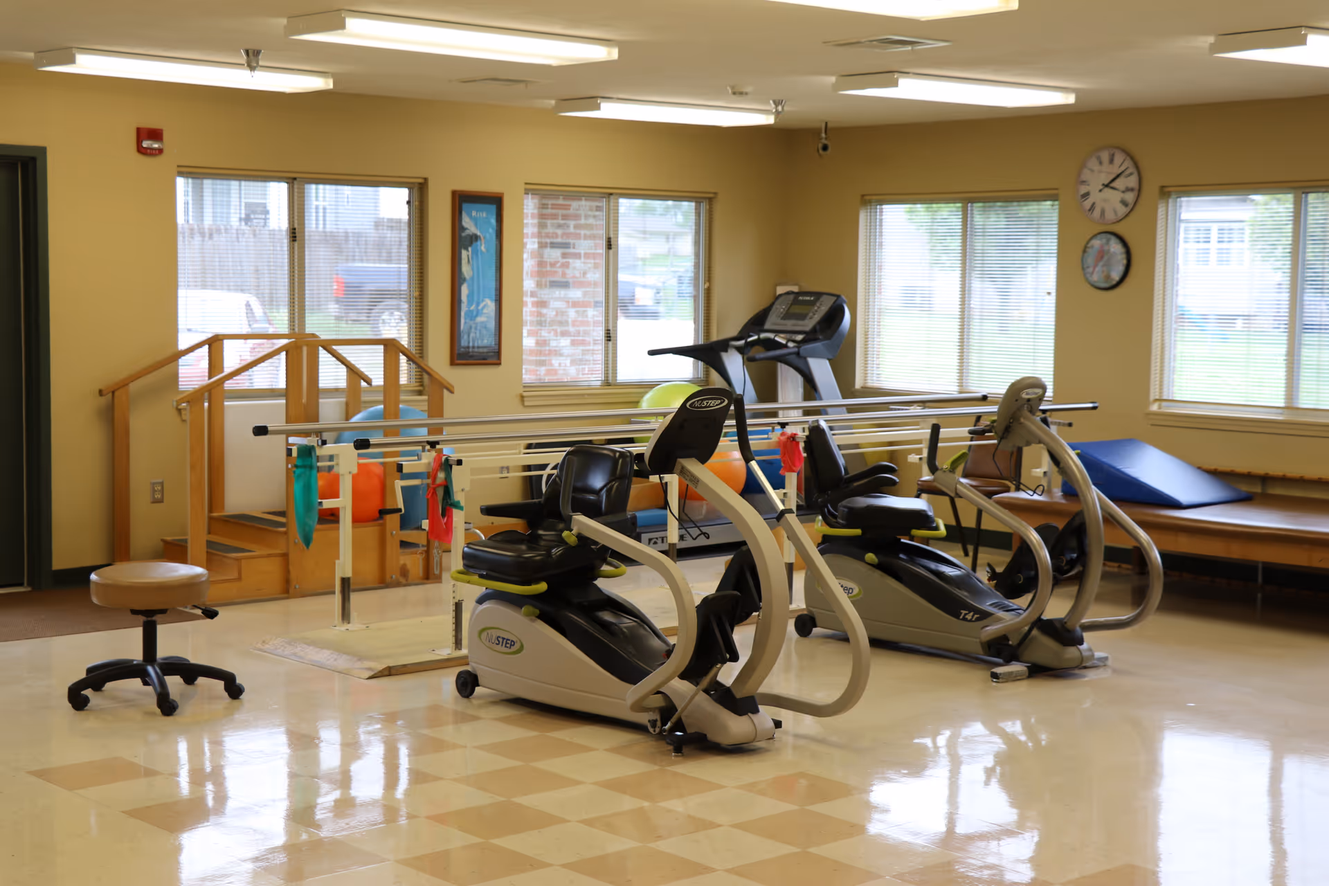A rehabilitation or physical therapy room with exercise equipment including two recumbent exercise bikes, parallel bars for walking practice, a treadmill, a small set of wooden stairs with handrails, and a padded bench with a blue wedge cushion. The room has large windows letting in natural light and a clock on the wall.