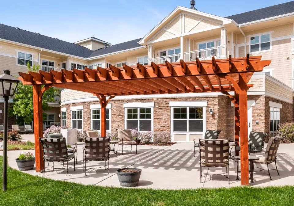 Outdoor courtyard with a wooden pergola shading patio chairs in front of a multi-story senior living building.