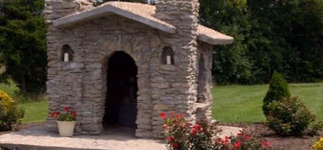 A small stone structure resembling a miniature house or chapel with an arched entrance and two small windows, surrounded by a well-maintained garden with green grass, bushes, and red flowers.