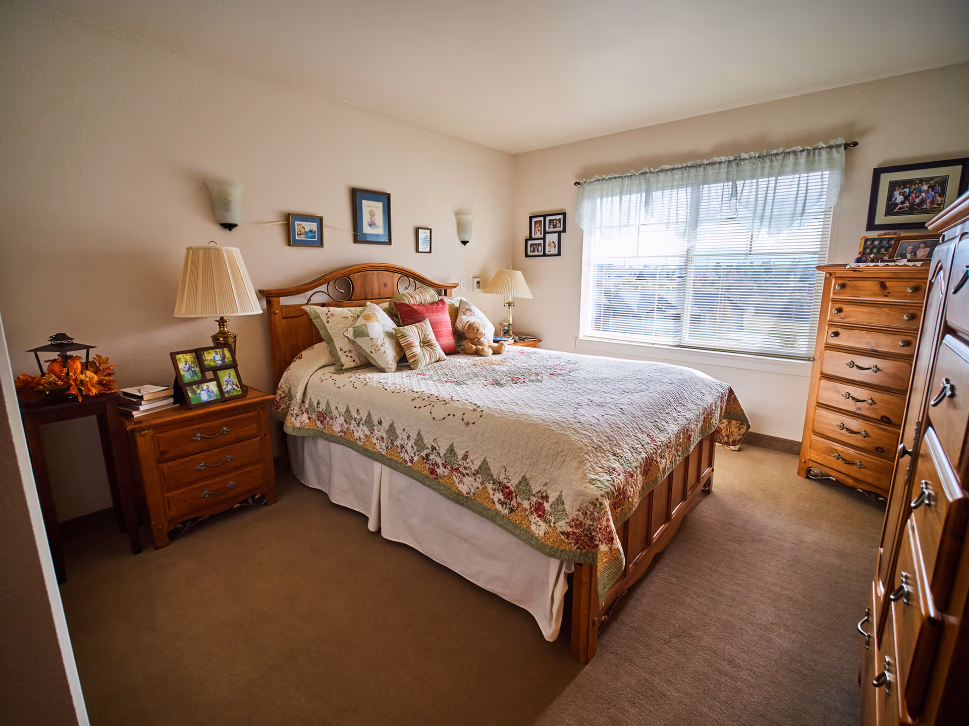 Bright bedroom with a wooden bed covered in a floral quilt, bedside tables, lamps, and dressers.
