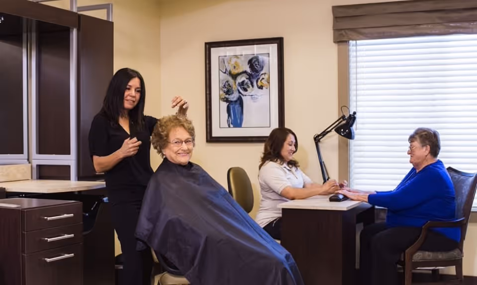 A senior woman is getting her hair styled by a hairdresser in a salon area, while another senior woman is receiving a manicure from a nail technician at a nearby table. The room has beige walls, a framed floral painting, and a window with blinds.