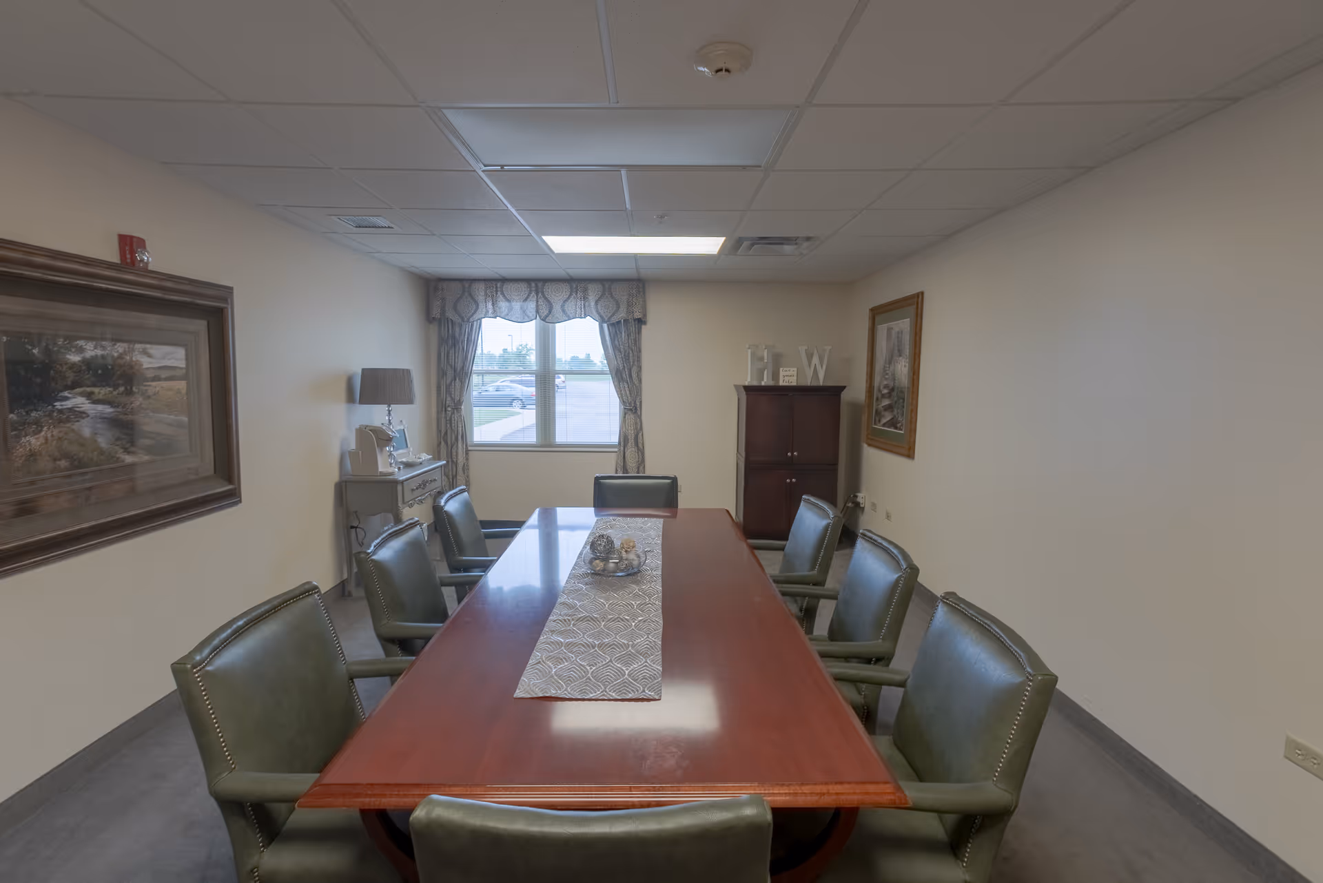 A conference or meeting room with a long wooden table surrounded by eight green upholstered chairs. The room has beige walls, a window with patterned curtains, a small cabinet with decorative items, a lamp on a side table, and framed artwork on the walls.