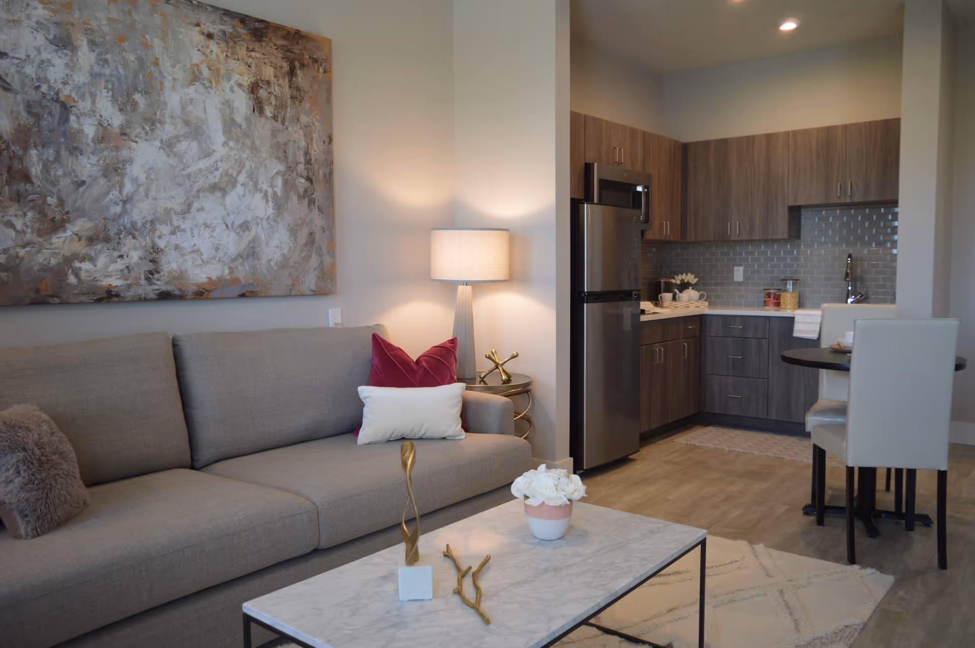 A modern living area with a gray sofa adorned with decorative pillows, a marble coffee table with decorative items, a side table with a lamp, and an abstract painting on the wall. In the background, there is a kitchen with wooden cabinets, a stainless steel refrigerator, microwave, and a small dining table with two white chairs.