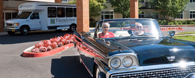 Two older adults sit in a vintage convertible near the entrance of Friendsview Retirement Community, with a Friendsview shuttle van and flowerbeds in the background.