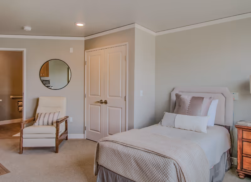 A tidy bedroom with a single bed, bedside table, armchair, round wall mirror, and double closet doors.