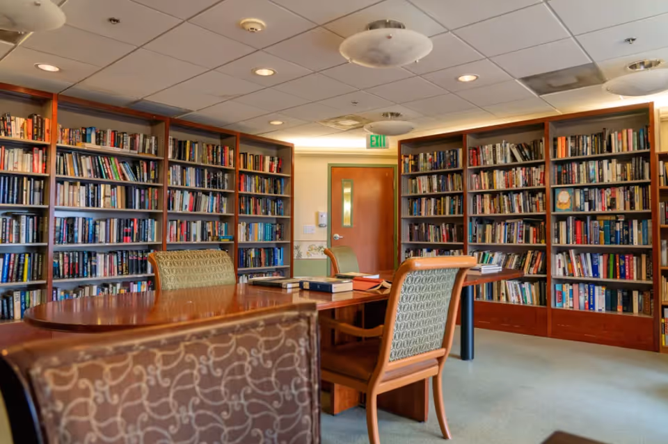 A quiet library room with wooden bookshelves filled with books lining the walls. There is a wooden table in the center with several chairs around it, some with patterned upholstery. The room has a drop ceiling with recessed lighting and an exit door in the background.