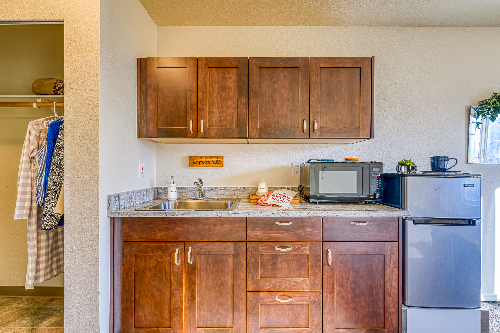 A small kitchenette area with wooden cabinets above and below a granite countertop. The countertop has a stainless steel sink, a soap dispenser, a microwave, a mini refrigerator, and some decorative items including a small plant and a mug. To the left, there is an open closet with clothes hanging inside. A small wooden sign on the wall reads 'Happiness is homemade.'
